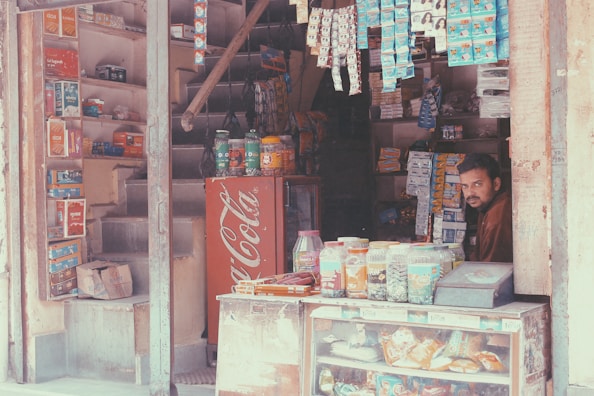 A small shop with shelves full of various packaged goods, including snacks and household items. There is a prominent Coca-Cola cooler in red with white text near the entrance. A man is sitting inside the shop, surrounded by hanging packages and jars filled with snacks.