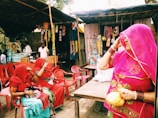 A street market scene features three women dressed in vibrant traditional attire with veils, sitting on plastic chairs. Various packaged snacks hang from a stall in the background, and a vendor stands behind a display of clay cups and bottled beverages.
