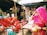 A street market scene features three women dressed in vibrant traditional attire with veils, sitting on plastic chairs. Various packaged snacks hang from a stall in the background, and a vendor stands behind a display of clay cups and bottled beverages.