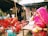 A street market scene features three women dressed in vibrant traditional attire with veils, sitting on plastic chairs. Various packaged snacks hang from a stall in the background, and a vendor stands behind a display of clay cups and bottled beverages.