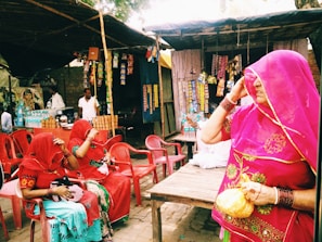 A street market scene features three women dressed in vibrant traditional attire with veils, sitting on plastic chairs. Various packaged snacks hang from a stall in the background, and a vendor stands behind a display of clay cups and bottled beverages.
