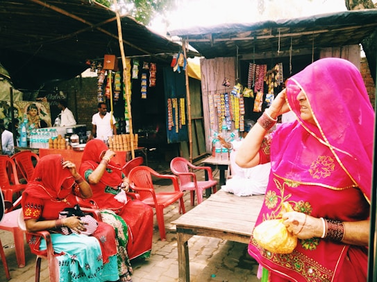 A street market scene features three women dressed in vibrant traditional attire with veils, sitting on plastic chairs. Various packaged snacks hang from a stall in the background, and a vendor stands behind a display of clay cups and bottled beverages.