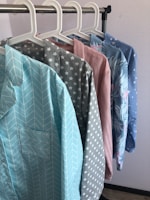 Several colorful shirts hang in a row on white hangers against a light-colored wall. Each shirt features a distinct pattern, including chevrons, stars, solid pink, floral designs, and small stars.