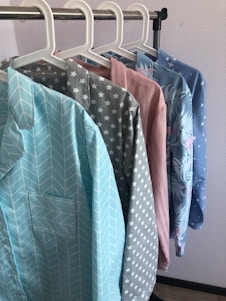 Several colorful shirts hang in a row on white hangers against a light-colored wall. Each shirt features a distinct pattern, including chevrons, stars, solid pink, floral designs, and small stars.