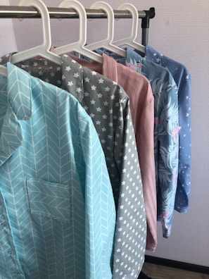 Several colorful shirts hang in a row on white hangers against a light-colored wall. Each shirt features a distinct pattern, including chevrons, stars, solid pink, floral designs, and small stars.