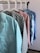 Several colorful shirts hang in a row on white hangers against a light-colored wall. Each shirt features a distinct pattern, including chevrons, stars, solid pink, floral designs, and small stars.
