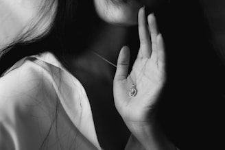 A close-up of a person's hand holding a delicate necklace with a round pendant featuring a textured design. The lighting is dramatic, creating deep shadows and highlights, adding a sense of mystery and elegance.