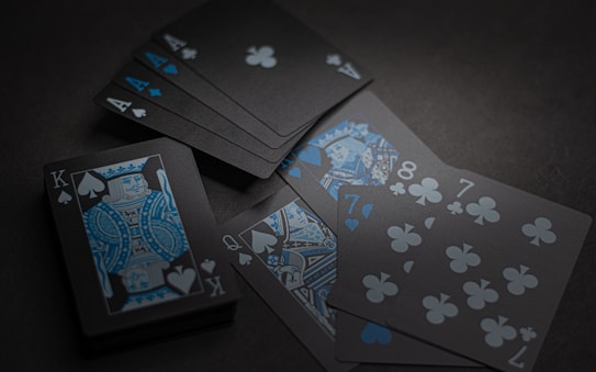 A set of playing cards with a dark and stylish design featuring black backgrounds and white and blue symbols. The cards include a King, Queen, and several numbered cards, all laid out on a dark surface.