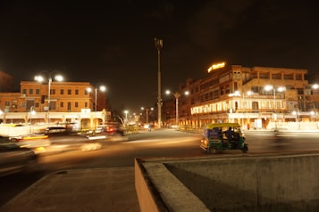 A nighttime urban scene with illuminated buildings displaying a traditional architectural style. The street is active with light trails from moving vehicles, and a green and yellow auto rickshaw is visible on the side of the road. Streetlights and building lights create a warm ambiance, contrasting against the dark sky.