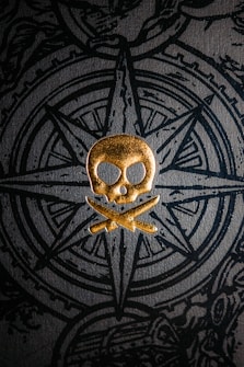 A golden skull and crossbones emblem is prominently displayed against a textured dark background featuring an intricate compass design. The design incorporates detailed line work and a sense of depth, creating a contrast between the gold and black elements.