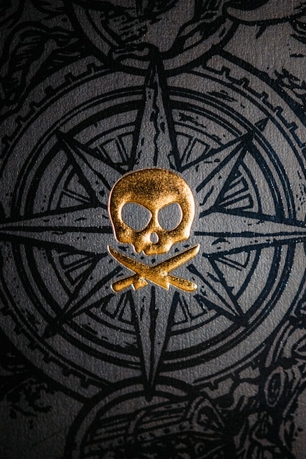 A golden skull and crossbones emblem is prominently displayed against a textured dark background featuring an intricate compass design. The design incorporates detailed line work and a sense of depth, creating a contrast between the gold and black elements.