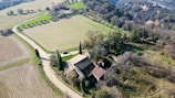 An aerial view of a rustic farmhouse surrounded by fields and rows of trees. The landscape includes a winding dirt road leading to the farmhouse, which is characterized by its terracotta roof and stone walls. In the background, dense forested hills provide a natural border.