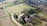 An aerial view of a rustic farmhouse surrounded by fields and rows of trees. The landscape includes a winding dirt road leading to the farmhouse, which is characterized by its terracotta roof and stone walls. In the background, dense forested hills provide a natural border.