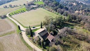 An aerial view of a rustic farmhouse surrounded by fields and rows of trees. The landscape includes a winding dirt road leading to the farmhouse, which is characterized by its terracotta roof and stone walls. In the background, dense forested hills provide a natural border.