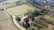 An aerial view of a rustic farmhouse surrounded by fields and rows of trees. The landscape includes a winding dirt road leading to the farmhouse, which is characterized by its terracotta roof and stone walls. In the background, dense forested hills provide a natural border.