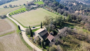 An aerial view of a rustic farmhouse surrounded by fields and rows of trees. The landscape includes a winding dirt road leading to the farmhouse, which is characterized by its terracotta roof and stone walls. In the background, dense forested hills provide a natural border.