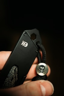 A close-up of a black knife being held by someone's fingers. The knife features a skull logo on its surface, and it has a black cord with a silver button that also has a skull design.