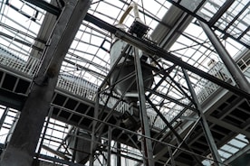 An industrial interior filled with metal beams and supports, showcasing a complex structure of girders and railings. The ceiling is comprised of glass panels allowing natural light to illuminate the space. The architectural design features intricate and heavy-duty construction elements.