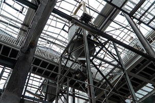An industrial interior filled with metal beams and supports, showcasing a complex structure of girders and railings. The ceiling is comprised of glass panels allowing natural light to illuminate the space. The architectural design features intricate and heavy-duty construction elements.