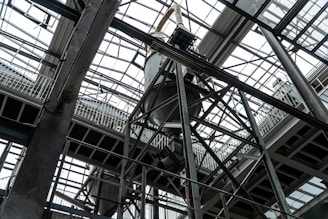 An industrial interior filled with metal beams and supports, showcasing a complex structure of girders and railings. The ceiling is comprised of glass panels allowing natural light to illuminate the space. The architectural design features intricate and heavy-duty construction elements.