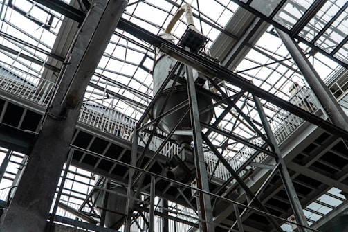 An industrial interior filled with metal beams and supports, showcasing a complex structure of girders and railings. The ceiling is comprised of glass panels allowing natural light to illuminate the space. The architectural design features intricate and heavy-duty construction elements.