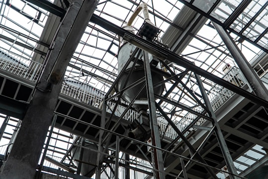 An industrial interior filled with metal beams and supports, showcasing a complex structure of girders and railings. The ceiling is comprised of glass panels allowing natural light to illuminate the space. The architectural design features intricate and heavy-duty construction elements.