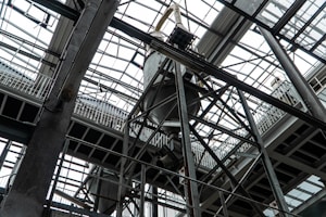 An industrial interior filled with metal beams and supports, showcasing a complex structure of girders and railings. The ceiling is comprised of glass panels allowing natural light to illuminate the space. The architectural design features intricate and heavy-duty construction elements.