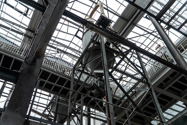 An industrial interior filled with metal beams and supports, showcasing a complex structure of girders and railings. The ceiling is comprised of glass panels allowing natural light to illuminate the space. The architectural design features intricate and heavy-duty construction elements.