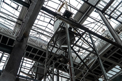 An industrial interior filled with metal beams and supports, showcasing a complex structure of girders and railings. The ceiling is comprised of glass panels allowing natural light to illuminate the space. The architectural design features intricate and heavy-duty construction elements.