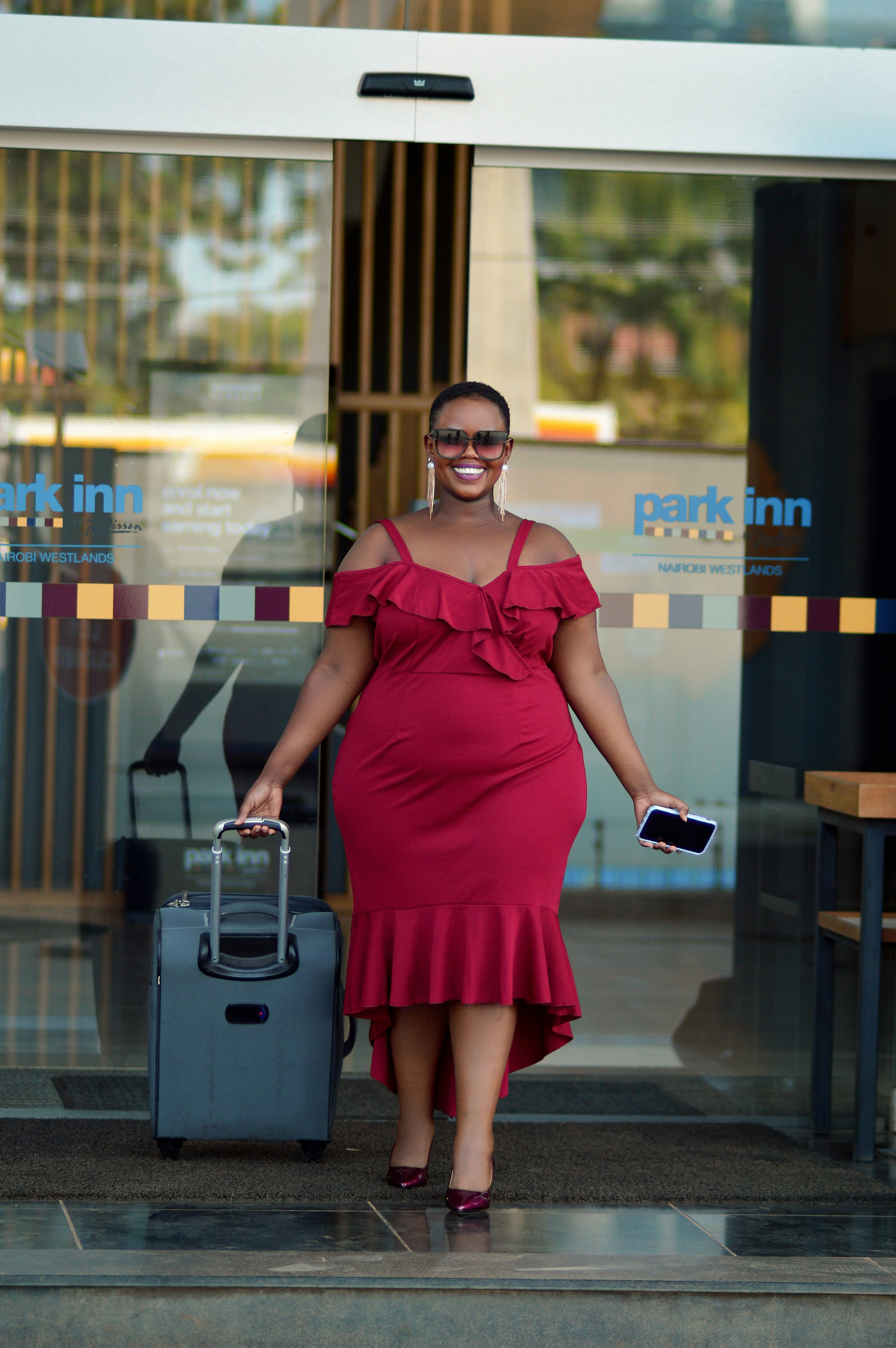 A person wearing a red dress and sunglasses is walking out of a hotel entrance with a suitcase in one hand and a phone in the other. The entrance features a glass door with the hotel logo 'park inn' visible.