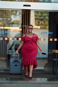 A person wearing a red dress and sunglasses is walking out of a hotel entrance with a suitcase in one hand and a phone in the other. The entrance features a glass door with the hotel logo 'park inn' visible.