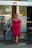 A person wearing a red dress and sunglasses is walking out of a hotel entrance with a suitcase in one hand and a phone in the other. The entrance features a glass door with the hotel logo 'park inn' visible.