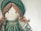 A fabric doll with an embroidered face that includes stitched eyes and a small smile. The doll has yarn hair styled in braids and wears a vintage-style outfit consisting of a green bonnet and dress with lace detailing.