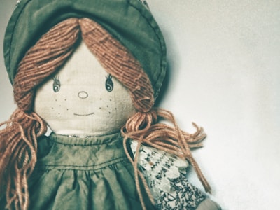 A fabric doll with an embroidered face that includes stitched eyes and a small smile. The doll has yarn hair styled in braids and wears a vintage-style outfit consisting of a green bonnet and dress with lace detailing.