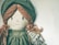 A fabric doll with an embroidered face that includes stitched eyes and a small smile. The doll has yarn hair styled in braids and wears a vintage-style outfit consisting of a green bonnet and dress with lace detailing.