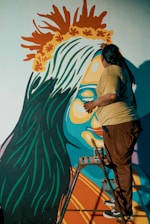 A person is on a stepladder painting a vibrant mural on a wall. The mural features a large, colorful figure with blue skin, orange and yellow facial features, and a crown-like headpiece made of orange and yellow leaves. The person's back is to the viewer, and they are adding details to the artwork.