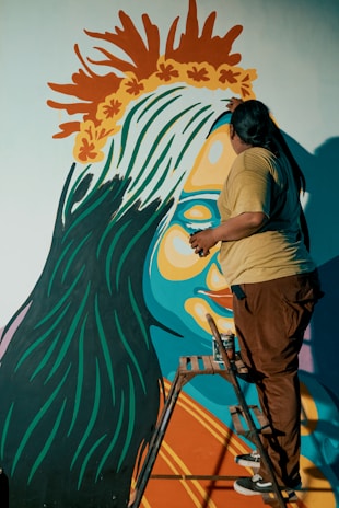 A person is on a stepladder painting a vibrant mural on a wall. The mural features a large, colorful figure with blue skin, orange and yellow facial features, and a crown-like headpiece made of orange and yellow leaves. The person's back is to the viewer, and they are adding details to the artwork.