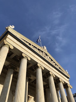A classical building featuring tall Ionic columns and intricate sculptures on the pediment. A Greek flag is prominently displayed atop the structure, set against a clear blue sky.