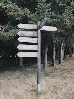 A wooden signpost with multiple directional arrows stands in a grassy area, surrounded by dense evergreen trees. The signpost has labels for various attractions such as boating, an elf and fairy village, an adventure course, archery, and a canopy cafe.