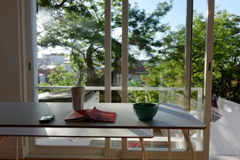 A bright room with a table set in front of a large window overlooking a garden. The table holds a smartphone, an open book titled 'Lisboa', a cup, and a green bowl with a spoon inside. Sunlight filters through the trees outside, casting shadows on the table.