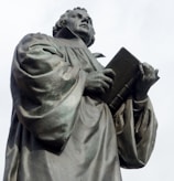 A bronze statue depicts a robed figure holding an open book and looking upwards with a thoughtful expression. The intricate folds of the robe and the detailed facial features convey a sense of reverence or scholarship.