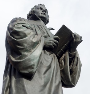 A bronze statue depicts a robed figure holding an open book and looking upwards with a thoughtful expression. The intricate folds of the robe and the detailed facial features convey a sense of reverence or scholarship.