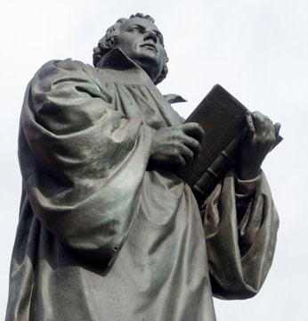 A bronze statue depicts a robed figure holding an open book and looking upwards with a thoughtful expression. The intricate folds of the robe and the detailed facial features convey a sense of reverence or scholarship.