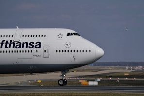 A large Lufthansa aircraft is parked on the runway. The front section of the plane, including its nose, cockpit, and part of the fuselage, is visible with the Lufthansa logo prominently displayed. In the background, a runway and another small aircraft are visible in the sky, indicating an airport setting. The scene has a clear sky with minimal cloud coverage.