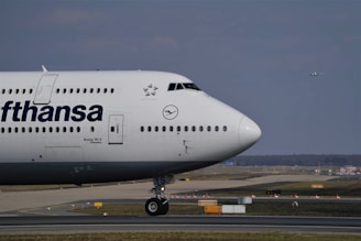 A large Lufthansa aircraft is parked on the runway. The front section of the plane, including its nose, cockpit, and part of the fuselage, is visible with the Lufthansa logo prominently displayed. In the background, a runway and another small aircraft are visible in the sky, indicating an airport setting. The scene has a clear sky with minimal cloud coverage.