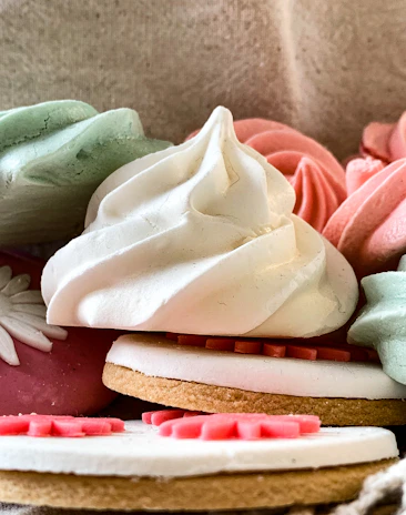 A close-up view of colorful, decorative meringue cookies and biscuits. The meringues are pastel-colored in shades of green, pink, and white, with intricate swirling patterns. One biscuit is visible with white icing and pink decorative elements on top.