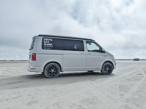 A white van is parked on a sandy beach with overcast skies. The side window has text advertisements or promotional statements. The background features open space with another vehicle in the distance.