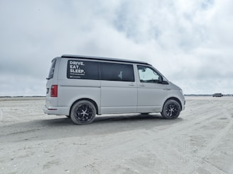 A white van is parked on a sandy beach with overcast skies. The side window has text advertisements or promotional statements. The background features open space with another vehicle in the distance.