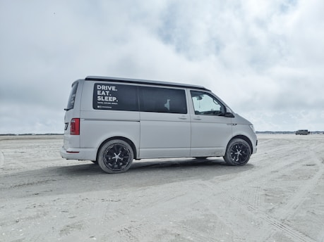A white van is parked on a sandy beach with overcast skies. The side window has text advertisements or promotional statements. The background features open space with another vehicle in the distance.