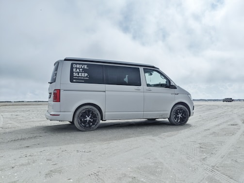 A white van is parked on a sandy beach with overcast skies. The side window has text advertisements or promotional statements. The background features open space with another vehicle in the distance.